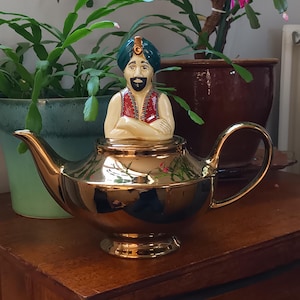 Puede incluir: Tetera dorada con una figura de genio. El genio lleva un turbante verde y un chaleco rojo. La tetera es reflectante y está sobre una superficie de madera. Objeto decorativo.