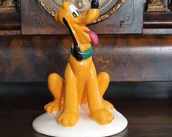 May include: A glossy ceramic figurine of the cartoon dog Pluto, featuring a bright yellow body, black ears, a green collar, and a pink tongue, sitting on a smooth white oval base with a cheerful expression and head tilted upward.