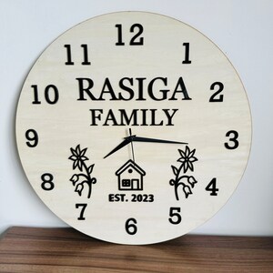 May include: A white wooden clock with black numbers and hands. The clock has the text "RASIGA FAMILY EST. 2023" on it. There are two stylized flower designs on either side of a small house design.