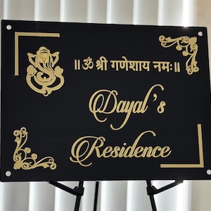 May include: Black rectangular sign with gold lettering and decorative accents. The sign features the name "Dayal's Residence" in elegant script, along with text in a different script and a gold illustration of a deity. The sign is mounted on a black stand.