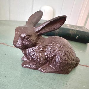 May include: A brown cast iron rabbit figurine with textured fur and long ears. The rabbit is sitting on a light green surface. The figurine is a decorative item, possibly for Easter or spring.