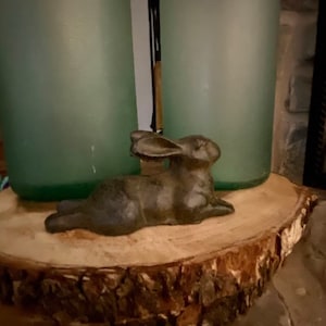 May include: A dark brown ceramic rabbit figurine lying on a wooden slice. The rabbit has long ears and is in a relaxed pose.