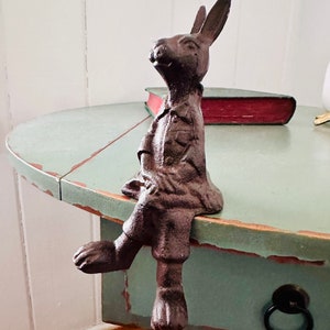 May include: A cast iron rabbit figurine, painted in a dark brown finish, sits on the edge of a light blue table. The rabbit is wearing a shirt and pants. A red book is visible in the background.