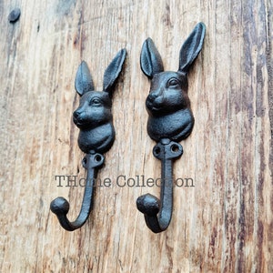 May include: Two black cast iron rabbit head hooks mounted on a wooden surface. The hooks have a simple design and are perfect for hanging coats, hats, or other items.