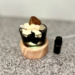 May include: A small, black glass bowl filled with various crystals and stones, resting on a wooden base with a warm light. A small, dark brown essential oil bottle sits to the side. The bowl contains a mix of white, brown, and black stones.