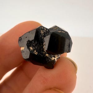 May include: A faceted, black tourmaline crystal with a glossy surface. The crystal has a dark, opaque appearance with some areas of lighter, beige-colored matrix. The mineral is held in a hand against a white background.