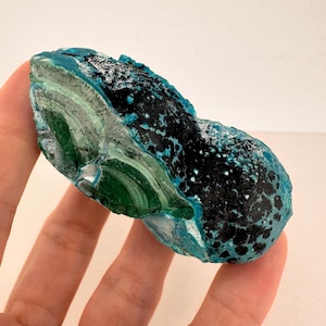 May include: A natural stone specimen displaying a rich palette of colours. The stone is predominantly teal, with patches of black and bands of vivid green. The irregular shape highlights the stone's natural, unrefined form, showcasing its unique character.
