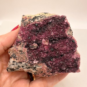 May include: A natural mineral specimen with a striking colour palette. The rock displays a deep magenta hue, contrasted by black areas and small green crystals. The texture is rough and crystalline, offering a unique visual appeal.