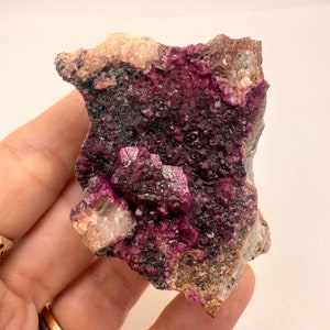 May include: A cluster of purple and black cobalt crystals on a matrix of grey rock. The crystals are iridescent and have a metallic sheen.