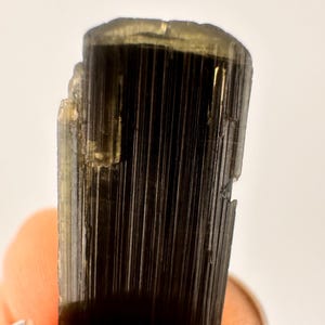 May include: Close-up of a dark, elongated crystal with vertical striations. The crystal has a deep black color with hints of gold and a slightly rough texture. The top edge is uneven, and the crystal is held by a person.