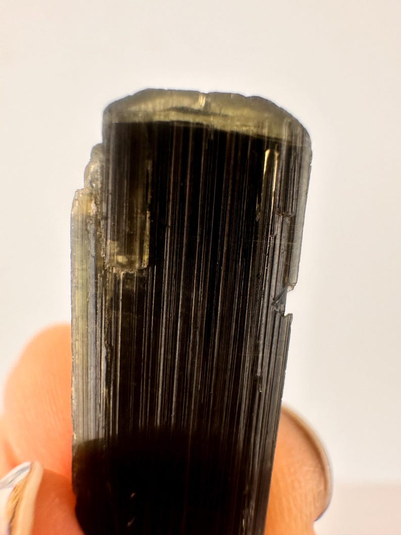 May include: Close-up of a dark, elongated crystal with vertical striations. The crystal has a deep black color with hints of gold and a slightly rough texture. The top edge is uneven, and the crystal is held by a person.