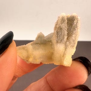 May include: A light yellow crystal cluster with a rough, textured surface. The crystal has a layered appearance with small, sparkling formations. The crystal is held between two fingers, showcasing its natural beauty and unique structure.