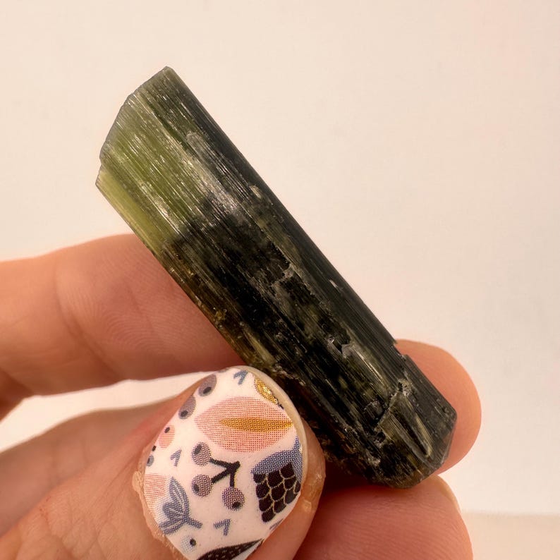 May include: A raw, elongated green tourmaline crystal. The gemstone has a dark green color with lighter green sections, and a natural, textured surface. The crystal is held against a white background.