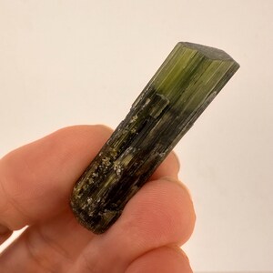 May include: A green tourmaline crystal with a dark, striated appearance. The gemstone is a long, rectangular shape with a faceted top. The crystal's color transitions from a deep green to a darker, almost black hue.