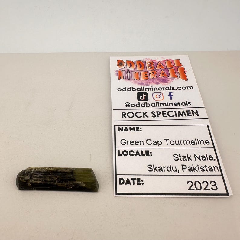 May include: A Green Cap Tourmaline rock specimen from Stak Nala, Skardu, Pakistan, dated 2023. The mineral is a dark green color with a lighter green cap. The specimen is next to a white card with the Oddball Minerals logo and details.