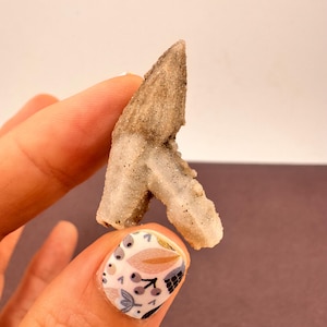 May include: A natural, light brown and white crystal formation with a textured surface. The crystal has a unique, angular shape. The crystal is a light beige colour with darker brown accents. It is held between fingers.