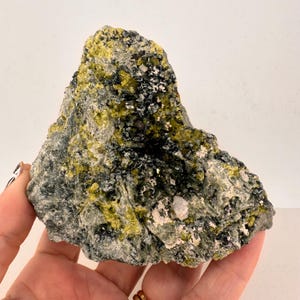 May include: A rough, gray rock with green and black crystals embedded in it. The rock has a natural, unpolished look.