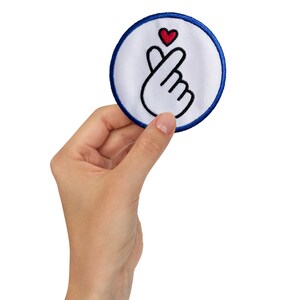 May include: A white fabric patch with a blue border, featuring a black outline of a hand making a heart shape with a red heart at the top.