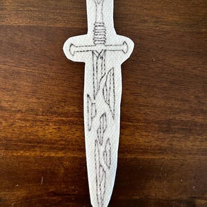 May include: A white felt sword with a black outline design. The sword features a detailed hilt, a crossguard, and a blade with a stylized pattern. The design is centered on a wooden surface.
