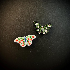 May include: Two butterfly-shaped pins. One is white with red and green floral designs, and a blue center. The other is black with white flowers and green leaves. Both pins have a glossy finish.