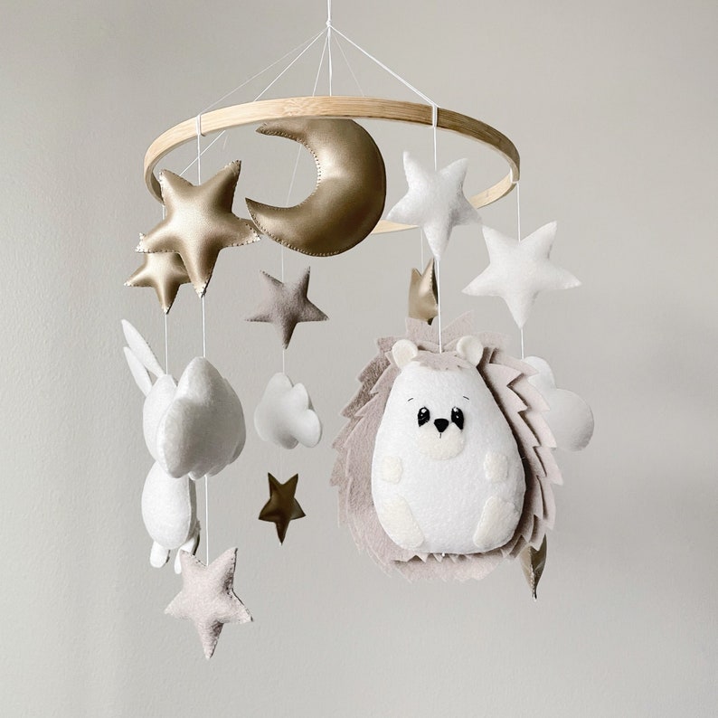 May include: A white and beige baby mobile with a wooden ring, a crescent moon, stars, clouds, a bunny, and a hedgehog.