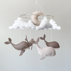 May include: A white and gray felt baby mobile with a crescent moon, clouds, and whales. The mobile is suspended from a gold metal ring.