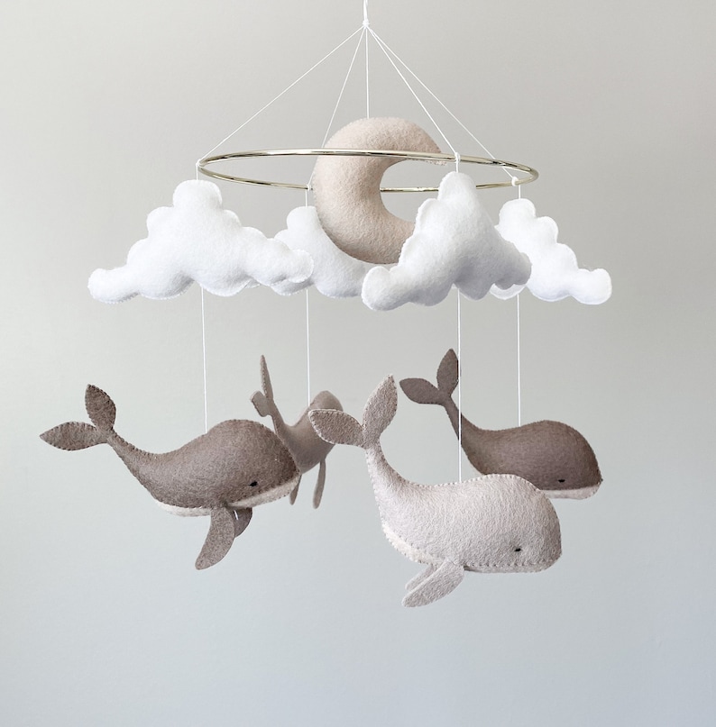 May include: A white and gray felt baby mobile with a crescent moon, clouds, and whales. The mobile is suspended from a gold metal ring.