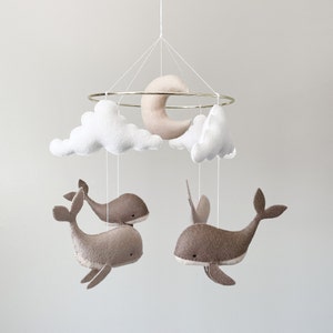 May include: A white and beige felt baby mobile with a crescent moon, clouds, and whales hanging from a gold metal ring.