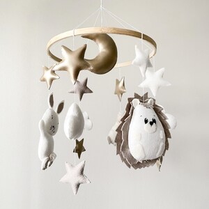 May include: A white and gold baby mobile with a crescent moon, stars, a rabbit, and a hedgehog. The mobile is made of felt and is hanging from a wooden ring.