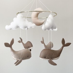 May include: A baby mobile with a gold metal ring, white clouds, a beige crescent moon, and four beige felt whales.