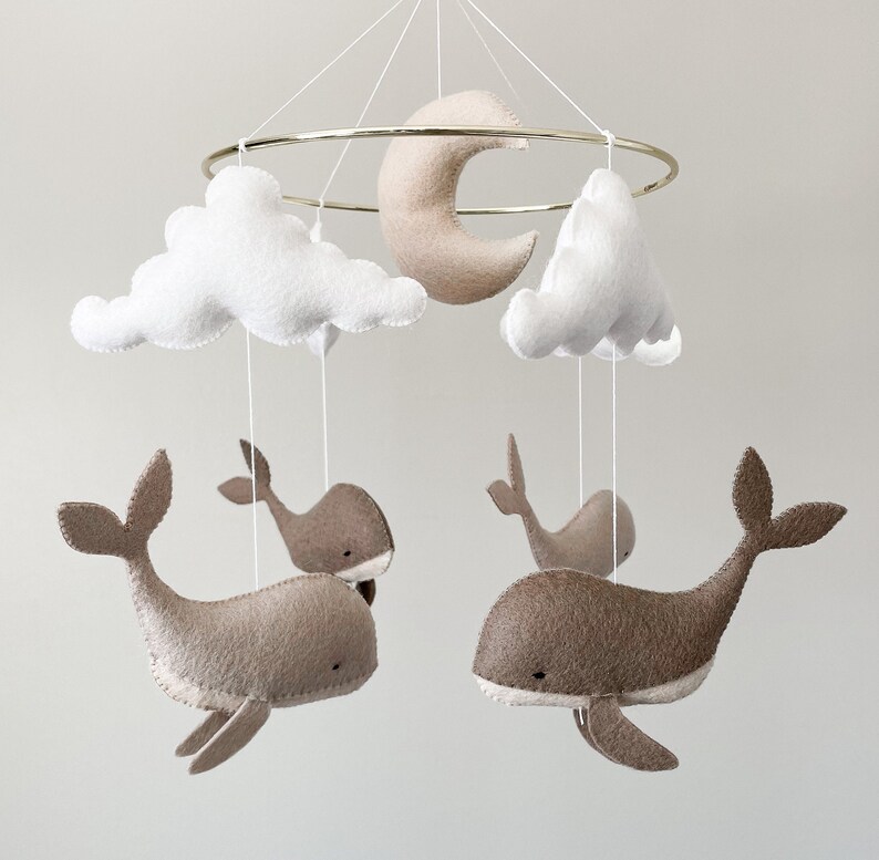 May include: A baby mobile with a gold metal ring, white clouds, a beige crescent moon, and four beige felt whales.