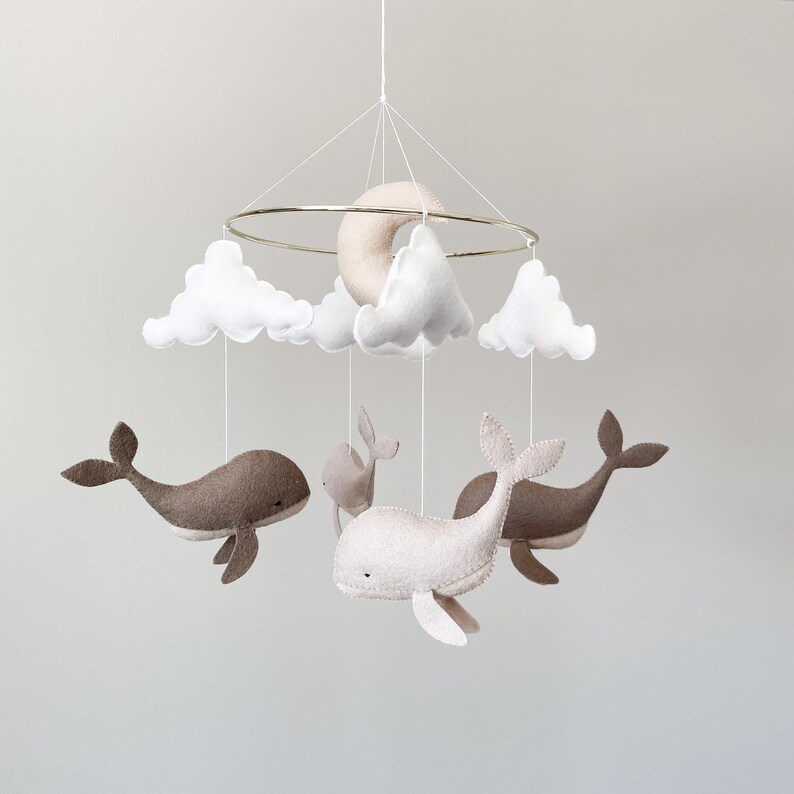 May include: A white and beige felt baby mobile with a crescent moon, clouds, and whales hanging from a gold ring.
