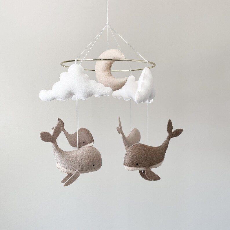 May include: A white and beige felt baby mobile with a gold metal ring. The mobile features two whales, a crescent moon, and two clouds.