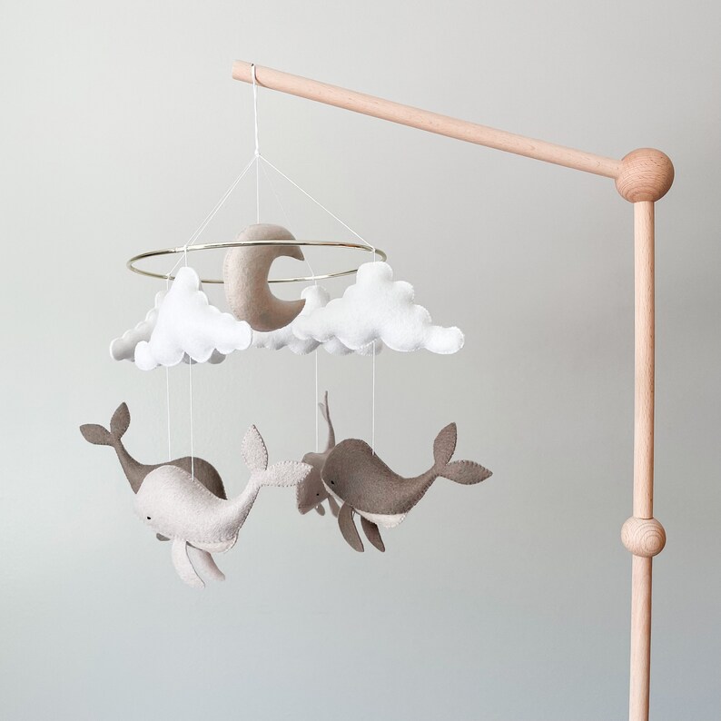 May include: A wooden mobile stand with a white and beige felt mobile hanging from it. The mobile features two whales, clouds, and a crescent moon.