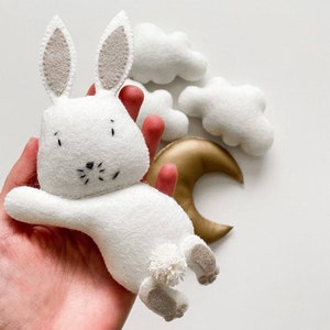 May include: A white felt bunny toy with a gold crescent moon and white felt clouds. The bunny has a fluffy tail and embroidered eyes and nose.