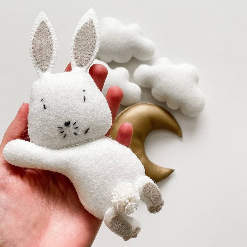 May include: A white felt bunny toy with a gold crescent moon and white felt clouds. The bunny has a fluffy tail and embroidered eyes and nose.