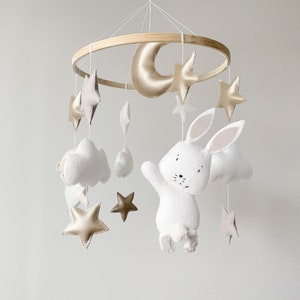 May include: A white felt bunny with a gold crescent moon, clouds, and stars hanging from a wooden ring. The mobile is a nursery decoration.
