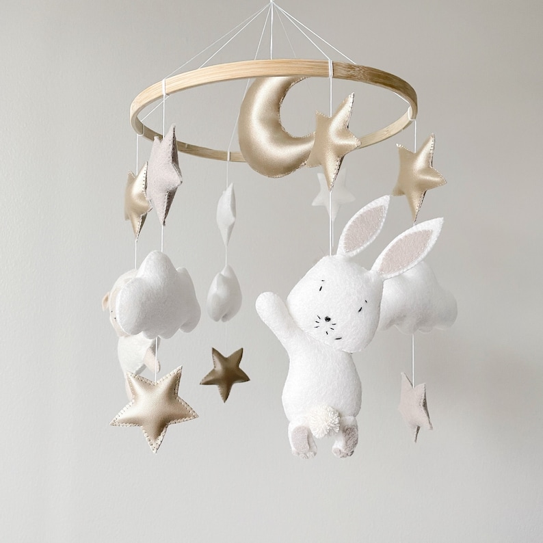 May include: A white felt bunny with a gold crescent moon, clouds, and stars hanging from a wooden ring. The mobile is a nursery decoration.