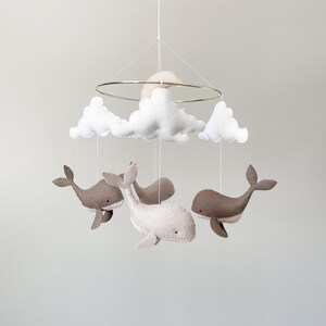 May include: A white and gray felt baby mobile with a crescent moon, clouds, and whales. The mobile is suspended from a gold metal ring.
