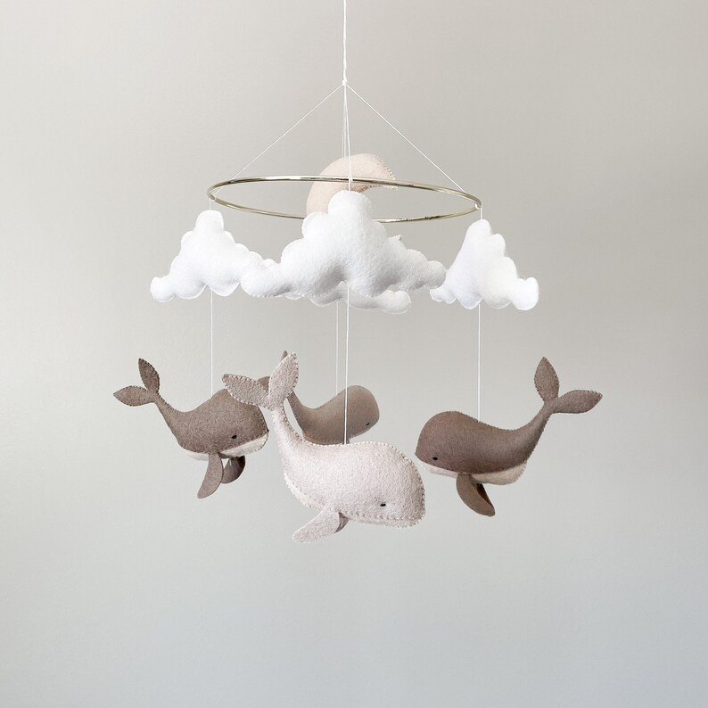 May include: A white and gray felt baby mobile with a crescent moon, clouds, and whales. The mobile is suspended from a gold metal ring.