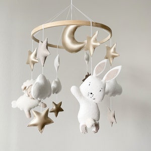 May include: A white felt bunny with a brown nose and black eyes hangs from a wooden mobile with gold stars, a crescent moon, and white clouds.