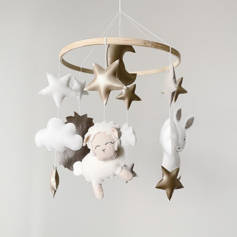 May include: A white and gold baby mobile with a lamb, clouds, stars, and a crescent moon hanging from a wooden ring.