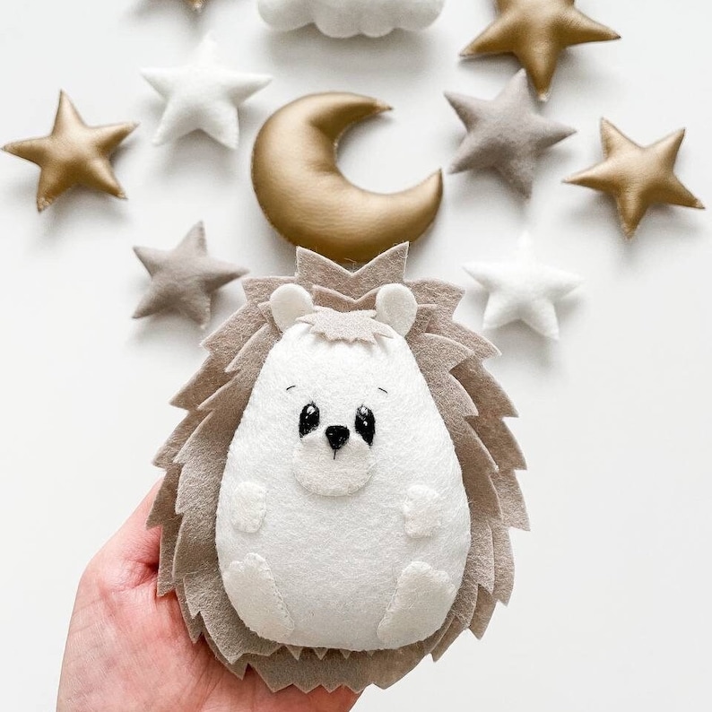 May include: A white felt hedgehog with black eyes and a brown felt body. The hedgehog is surrounded by felt stars and a crescent moon.