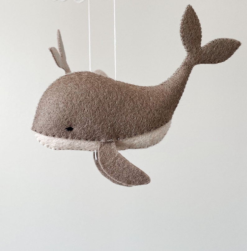 May include: A brown felt whale mobile hanging from a white string. The whale has a white felt belly and a black eye.