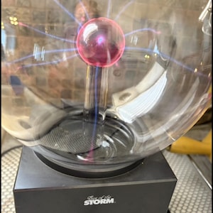 May include: A plasma ball lamp with a clear glass sphere and a pink central orb. Blue electrical arcs emanate from the center. The base is black and has the text "Eye of the Storm."