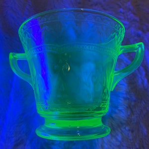 May include: A green glass sugar bowl with a decorative pattern and two handles. The bowl is illuminated with a blue light, making the green glass glow.