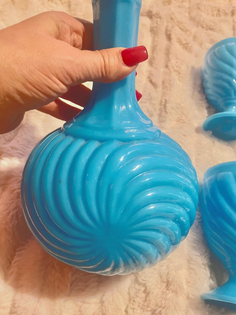 May include: A set of blue glass decanter and glasses with a swirl design. The decanter has a tall, slender neck and a round body. The glasses are shorter and have a wider bowl.