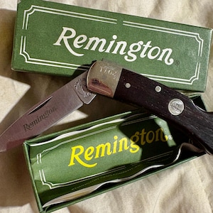 May include: A folding pocket knife with a dark wood handle and a silver blade, displayed with its green box. The box and blade are marked with the name "Remington". The knife is partially open, revealing the blade.