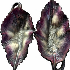 May include: Two leaf-shaped decorative dishes featuring a glossy finish with blended shades of deep purple, cream, and dark gray. Each dish has a textured surface, a serrated edge, and a small circular handle integrated into the stem area.