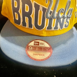 May include: A yellow and blue baseball cap with the words "UCLA BRUINS" embroidered on the front. The cap has a New Era logo and the words "9FIFTY SNAPBACK" on the back.
